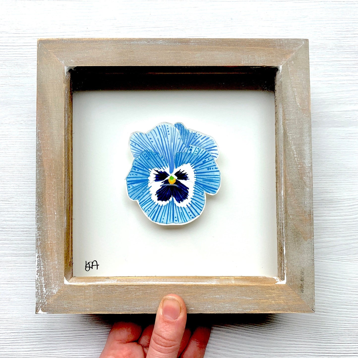 Framed artwork of a light blue and white pansy flower with a hand holding it on a white background