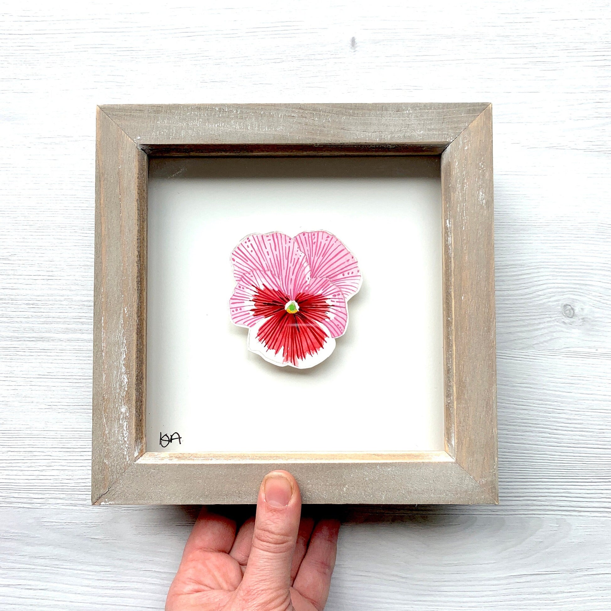 Hand holding a small wooden frame with a pink pansy flower painting on a white paper