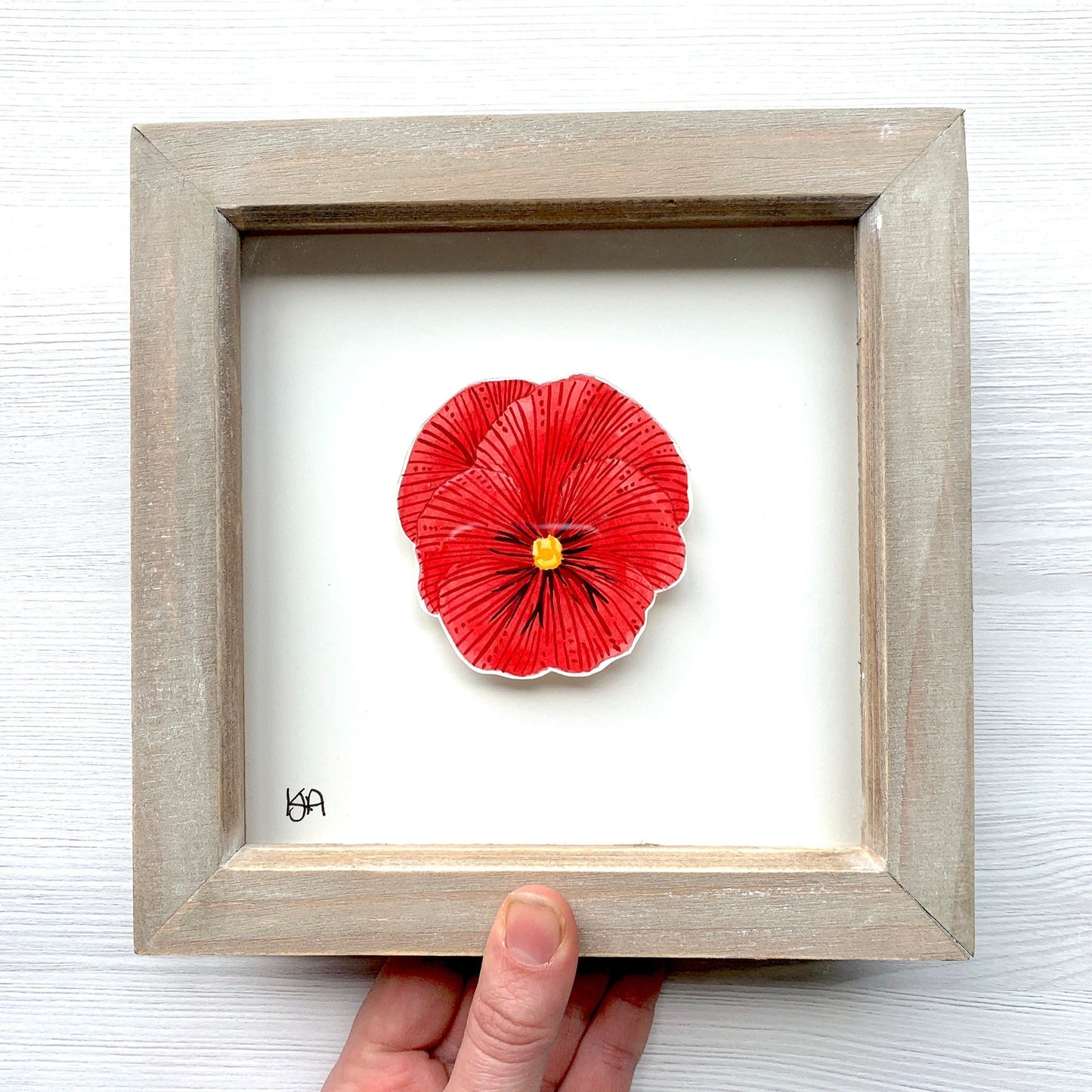 Framed artwork of a red pansy flower with a hand holding it against a white background