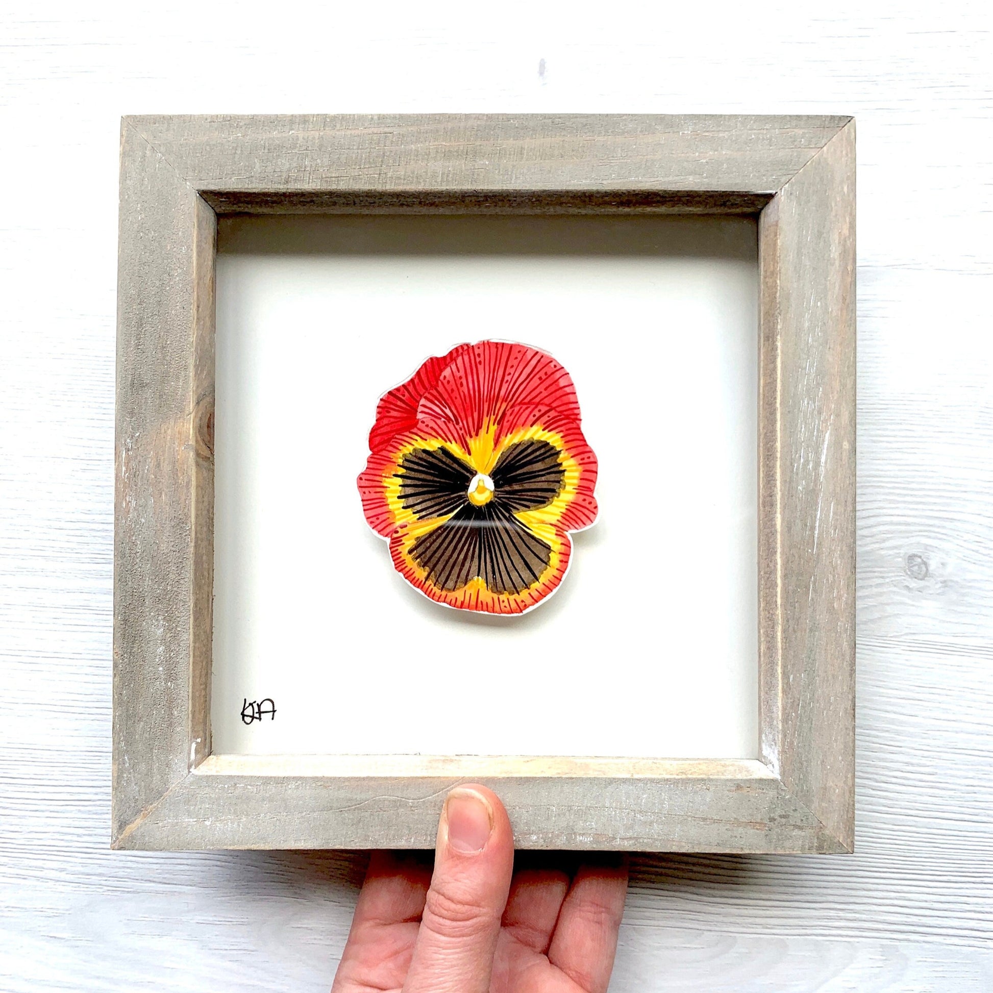 Framed artwork of a red, yellow and black pansy flower artwork in a frame held by a hand on a white background