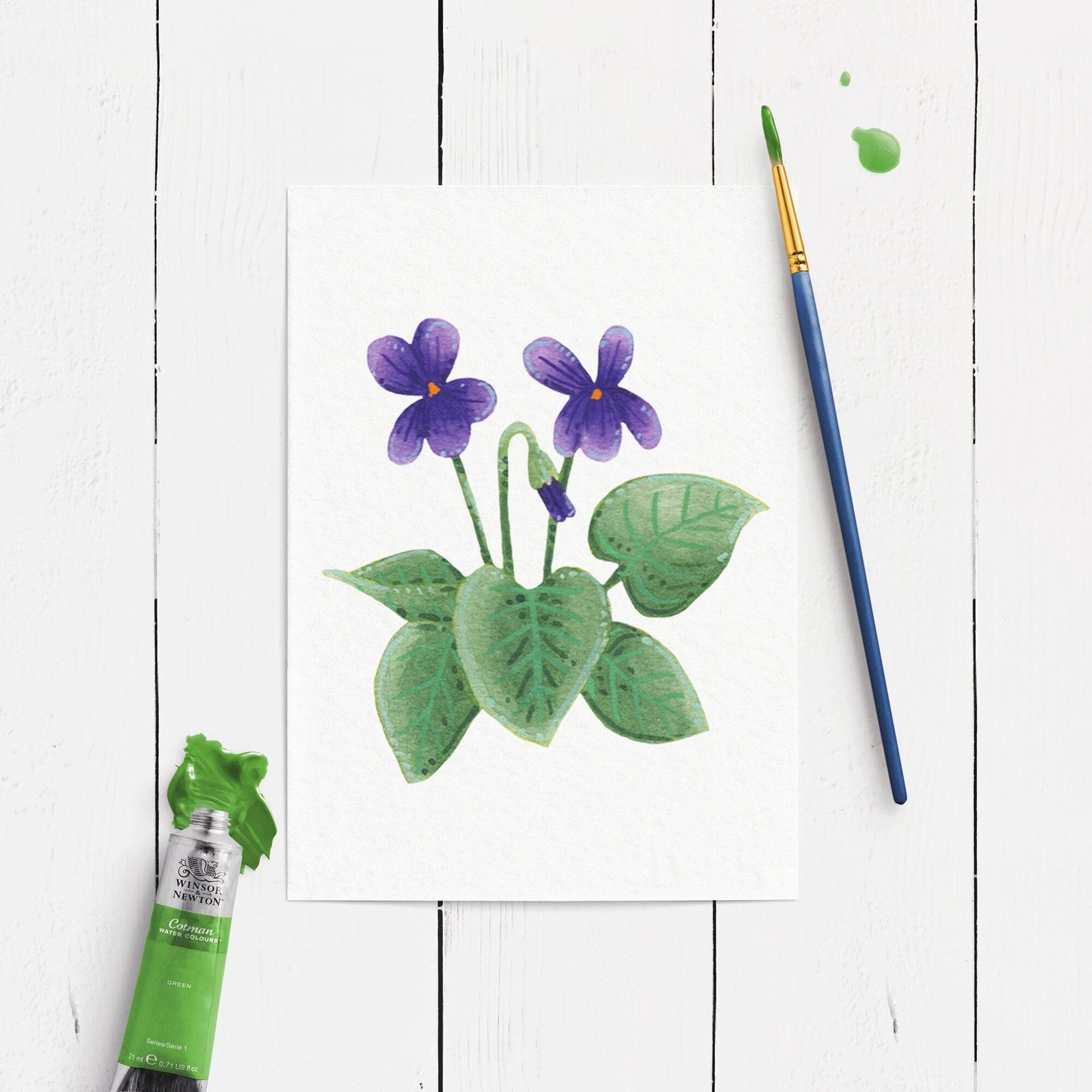 Painting of purple flowers with green leaves on a white wooden surface, accompanied by a paintbrush and paint tube.
