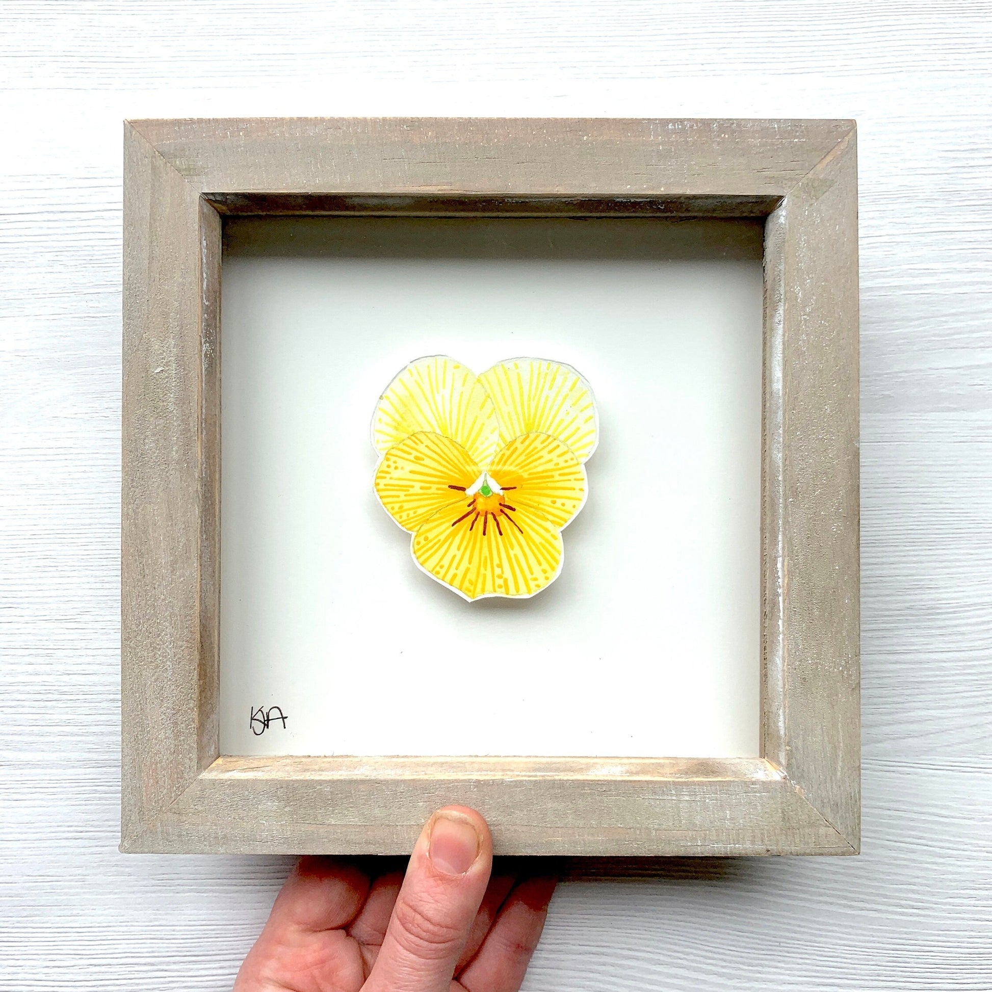 Yellow flower painting in a wooden frame held by a hand on a light wood background